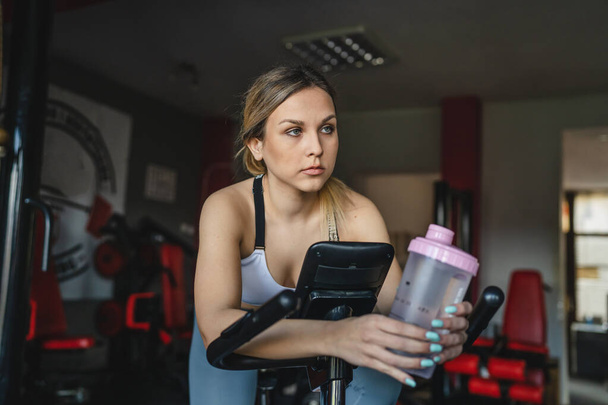 One woman at the gym training hold supplement shaker while riding spinning bike during workout real people caucasian female endurance and healthy life concept supplementation drink copy space - Foto, Bild
