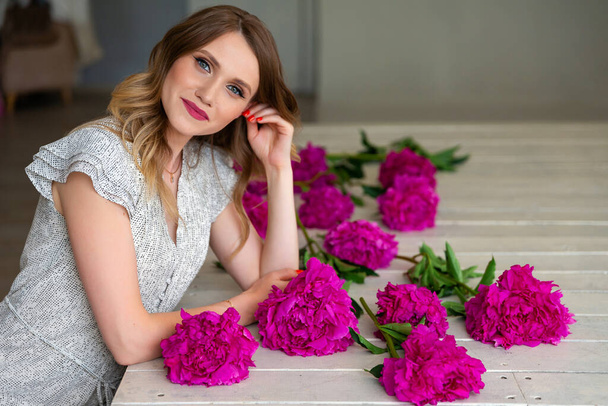 young woman in short dress sitting on wooden floor, near flowers of pink peonies. Place for your text. Concept of beauty and youth. Florist and designer worker - Fotó, kép