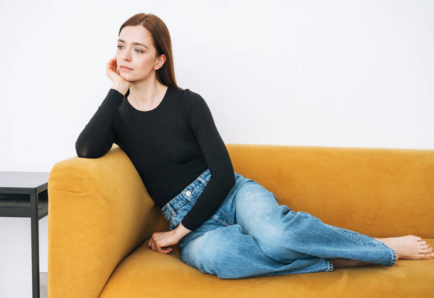 Young thinking woman girl with dark long hair in jeans sitting on yellow sofa in minimalistic interior - Foto, Imagem
