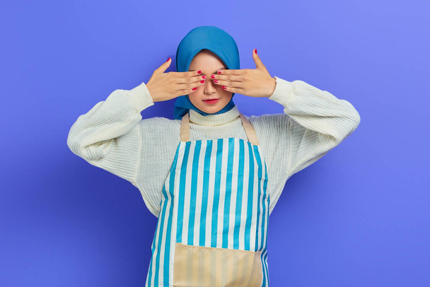 Krásná mladá žena v domácnosti v hidžábu a zástěra zakrývající oči s rukama nechce vypadat izolovaně na fialovém pozadí. Lidé hospodyňky muslimský životní styl koncept - Fotografie, Obrázek
