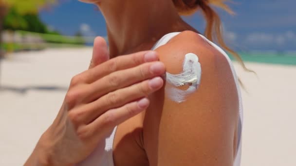 Close-up van een vrolijke glimlachende jonge vrouw is het aanbrengen van een zonnebrandcrème of zonnecrème op een schouder om te zorgen voor haar huid op een strand aan zee tijdens de vakantie. - Video