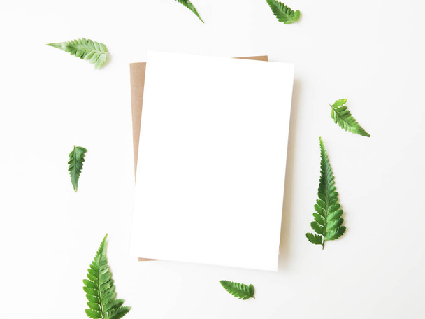 Blank greeting card mockup with kraft envelope surrounded by fern leaves on white table. Minimalist summer scene. Top view, flatlay, white copy space.  - Photo, Image