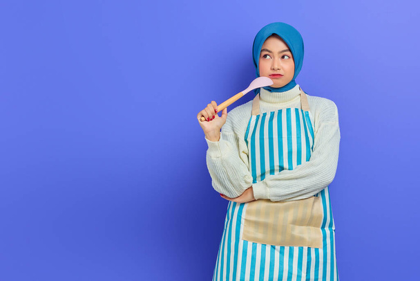 Pensive beautiful Asian woman in white sweater covered in apron and hijab holding a spoon, looking up doing housework isolated over purple background. Housekeeping concept - Fotoğraf, Görsel