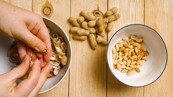 Top view of women's fingers peeling peanuts from their shells. A handful of peanuts lying on a wooden table. Peeling an earthy peanut at home - Foto, Imagem