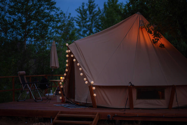 Tent on the territory of glamping at dusk. Lanterns glow on a tent in a campsite - Photo, Image