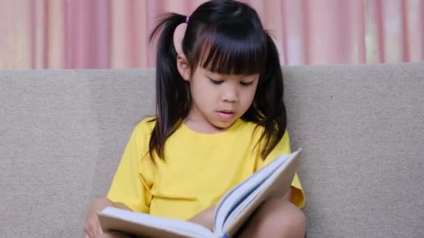 Linda niña leyendo un libro mientras está sentada en el sofá en casa. - Imágenes, Vídeo