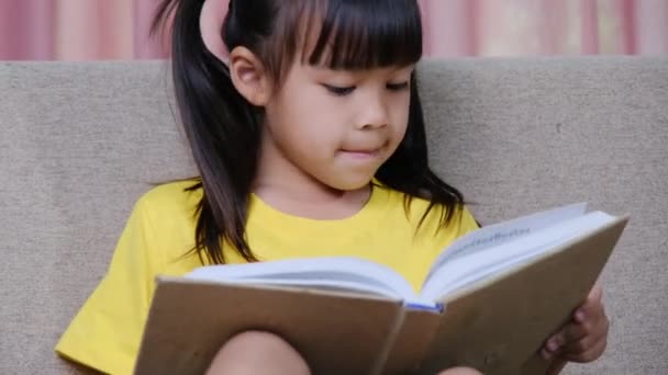 Söpö pieni tyttö lukee kirjaa istuessaan sohvalla kotona. - Materiaali, video