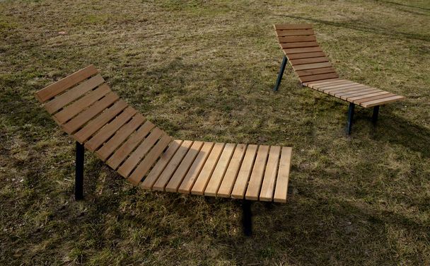 krzesło bujane na chodniku pod drzewami na placu. drewniane leżaki z drewna tropikalnego dla jednej osoby w parku. Są one komfortowo wykonane z brązowych desek, listew i stalowych ram - Zdjęcie, obraz