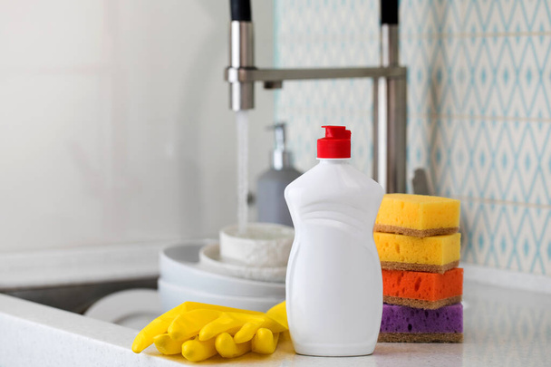 A white bottle with dishwashing gel, sponges and rubber gloves on the background of a sink with dirty dishes. Copy space - Foto, immagini