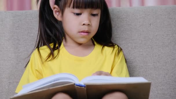 Cute little girl czytając książkę podczas siedzenia na kanapie w domu. - Materiał filmowy, wideo
