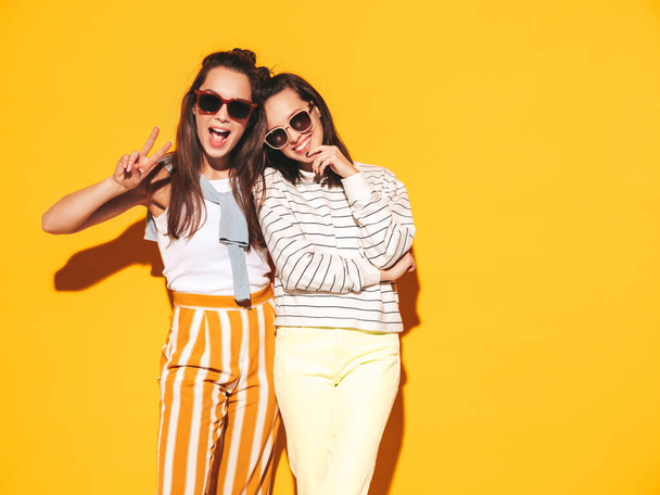 Two young beautiful smiling blond hipster female in trendy summer clothes. Sexy carefree women posing near yellow wall in studio. Positive models having fun. Cheerful and happy. In sunglasses - Foto, Imagem
