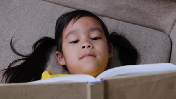 Aranyos kislány könyvet olvas, miközben otthon ül a kanapén.. - Felvétel, videó