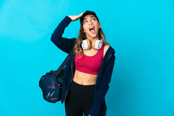 Young sport woman with sport bag isolated on blue background doing surprise gesture while looking to the side - Φωτογραφία, εικόνα