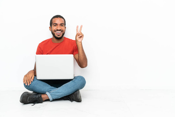 Mladý ekvádorský muž s laptopem, který sedí na podlaze izolovaný na bílém pozadí a usmívá se a ukazuje znamení vítězství - Fotografie, Obrázek