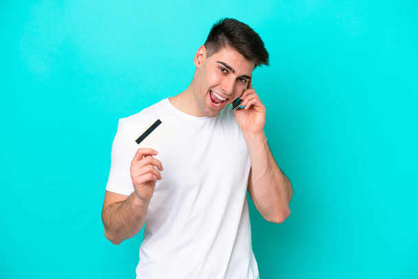 Joven hombre caucásico aislado sobre fondo azul manteniendo una conversación con el teléfono móvil y sosteniendo una tarjeta de crédito - Foto, Imagen