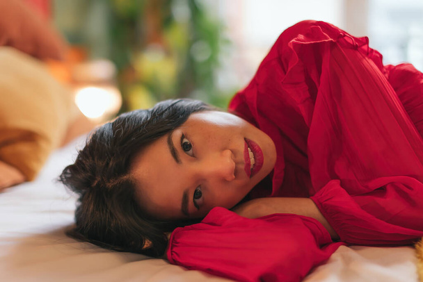 zblízka portrét mladé asijské ženy ležící na posteli s podmanivým a smyslným pohledem do kamery - intimní portrét ženy s rtěnkou a červenými šaty - Fotografie, Obrázek