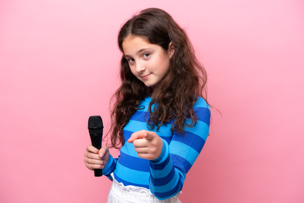 Pembe arka planda izole edilmiş bir mikrofonu eline alan küçük şarkıcı kız kendine güvenen bir ifadeyle sana parmak kaldırıyor. - Fotoğraf, Görsel