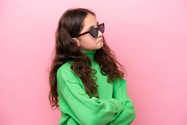Little caucasian girl wearing sunglasses isolated on pink background making doubts gesture while lifting the shoulders - Foto, afbeelding