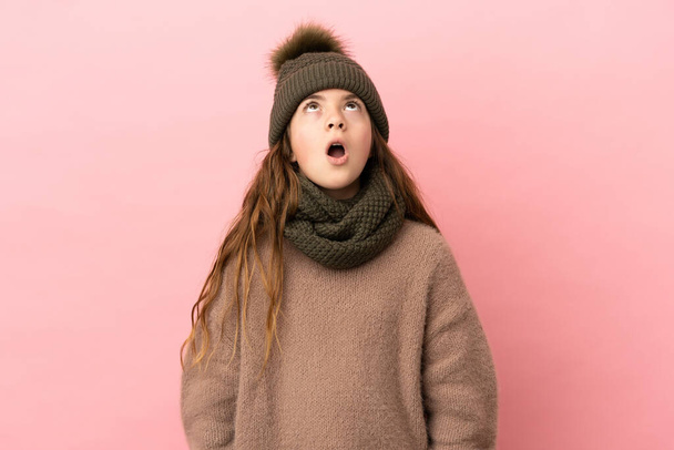 Little girl with winter hat isolated on pink background looking up and with surprised expression - Fotografie, Obrázek