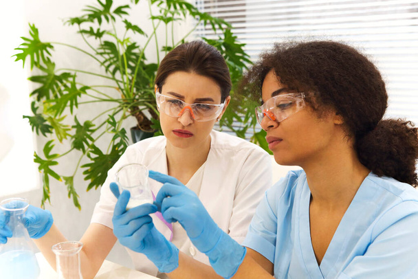 Testovací laboratoř COVID-19. Dvě ženy lékařských laboratoří kolegové nosí brýle pracující s zkumavkami, multi-etnický absolventi studenti pracují na výzkumném projektu - Fotografie, Obrázek