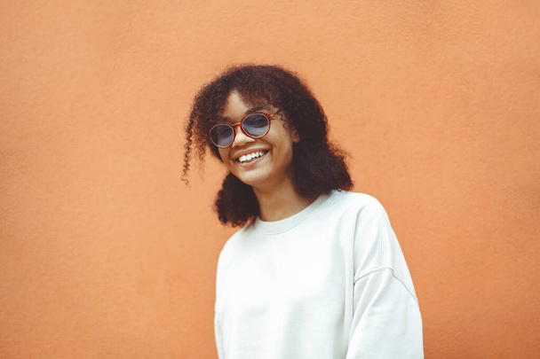 Mujer joven de etnia africana con gafas de sol elegantes, con el pelo rizado atado en cola de caballo alta, mirando hacia otro lado mientras sonríe ampliamente mostrando dientes rectos perfectos, posando sobre fondo de pared naranja - Foto, imagen