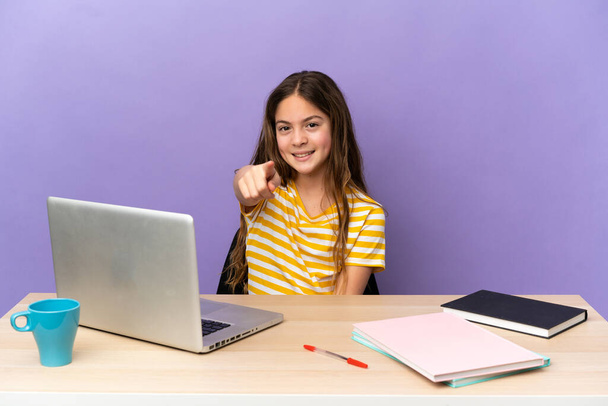 Kleines Studentenmädchen an einem Arbeitsplatz mit einem Laptop auf violettem Hintergrund, der nach vorne zeigt, mit glücklichem Gesichtsausdruck - Foto, Bild