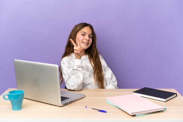 Mor arka planda dizüstü bilgisayarı olan küçük bir öğrenci kız gülümsüyor ve zafer işareti gösteriyor. - Fotoğraf, Görsel