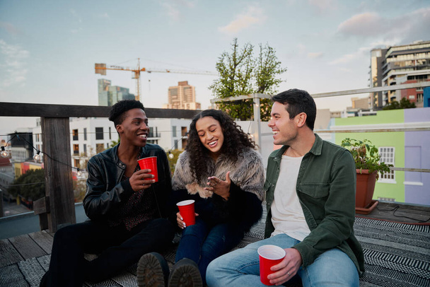 Gruppe multikultureller junger Erwachsener, die gemeinsam Witze lachen. Auf einer Dachparty in der Stadt sitzen, gute Zeiten. - Foto, Bild