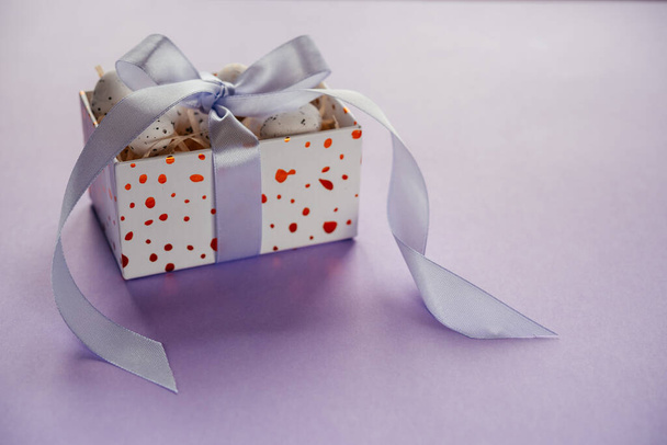 Пасхальный креативный макет с пасхальными яйцами в подарочной коробке с лавандовыми лентами румян на бледно-фиолетовом фоне - Фото, изображение