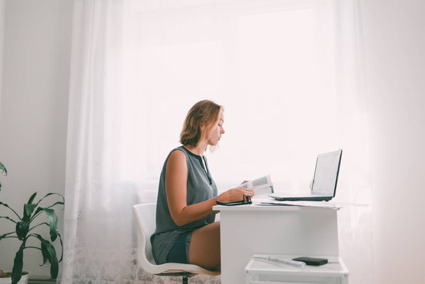 Młoda kobieta siedząca przy stole i przeglądająca książkę. W pobliżu znajduje się laptop i materiały biurowe i roślin. Wnętrze jest koloru białego. Przestrzeń kopiowania. - Zdjęcie, obraz