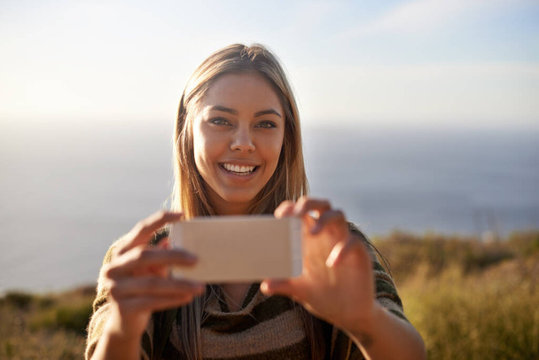 Un momento digno de recordar. Retrato de una atractiva joven sosteniendo su celular para tomar una foto al aire libre. - Foto, imagen