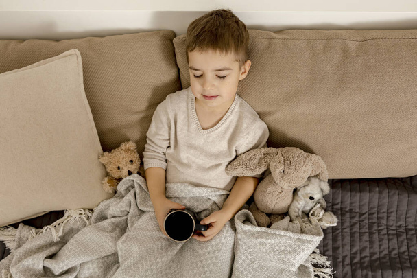 Очаровательный маленький мальчик пьет горячий чай на кровати дома и расслабляется. Ребенок отдыхает, завернутый в одеяло, с кружкой в комнате. Интерьер и одежда в натуральных цветах земли. Уютная обстановка. - Фото, изображение