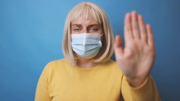 Młoda kobieta po trzydziestce w ochronnej masce medycznej z żółtym swetrem pokazującym gest zatrzymania na odizolowanym niebieskim tle - Materiał filmowy, wideo