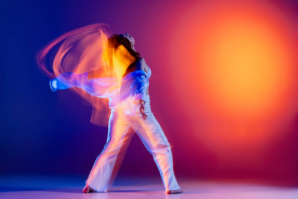 Image of flexible young girl, hip-hop dancer in white outfit dancing hip hop isolated on blue background in yellow neon light. Youth culture, style and fashion, action. - Photo, Image