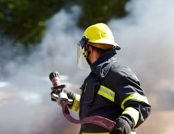 Hij heeft de situatie onder controle. Een Kaukasische brandweerman die een slang vasthield en omringd was door de rook van de brand.. - Foto, afbeelding