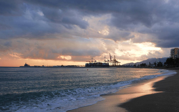Farbenfroher Sonnenuntergang über dem Seehafen und Industriekränen. Strandlinie mit Blick auf den Hafen von Spanien. Transport und logistischer Hintergrund - Seecontainer werden von Schiffen mit riesigen Kränen abtransportiert - Foto, Bild