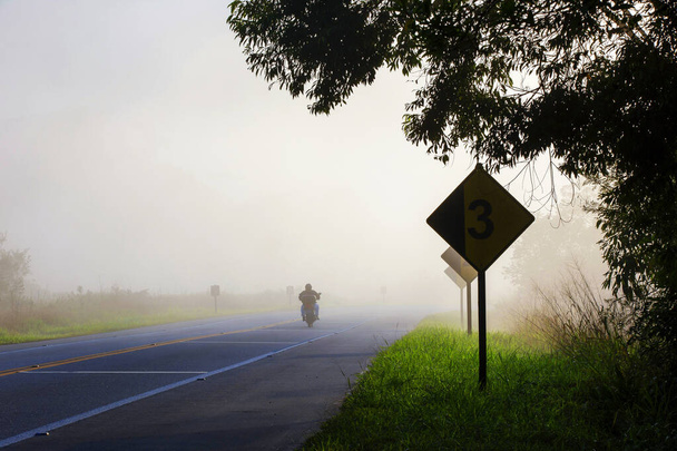 Auto-estrada na neblina da manhã, com motocicleta e sinais de alerta de cuidados. Rio Santos Road, São Paulo, Brasil
 - Foto, Imagem