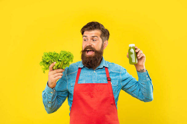 Happy man in apron looking at fresh lettuce holding juice bottle yellow background, juice barman - Foto, imagen