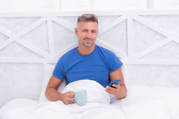 Happy man texting sms in smartphone while relaxing in bed with morning coffee ot tea, messaging - Фото, зображення