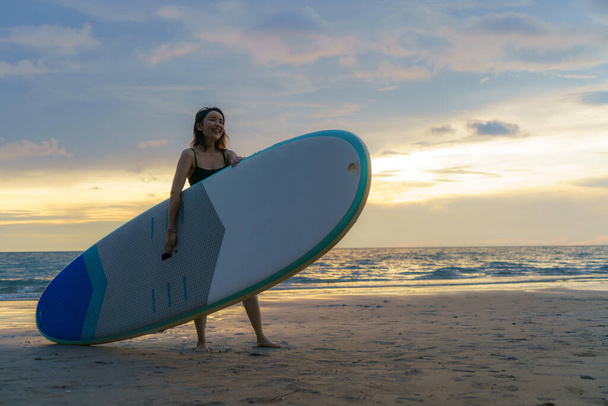 Mujer asiática sosteniendo una tabla de sup y remo y caminando por la playa. Al fondo, el océano y la puesta de sol. Vista trasera. Surf de verano. - Foto, Imagen