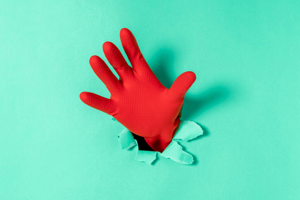 Hand in red rubber glove sticks out of a torn hole in blue paper - Zdjęcie, obraz