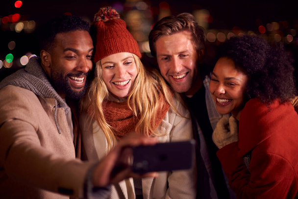 Group Of Friends Outdoors Wearing Coats And Scarves Posing For Selfie On Phone Against City Lights - Foto, Imagem