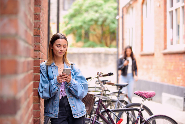 Young Woman Standing By Wall Looking At Mobile Phone Checking Text Messages And Social Media - Фото, изображение