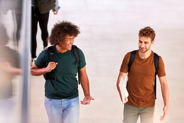 Two Male Students In Busy University Or College Building Talking As They Walk Along Corridor - Foto, afbeelding