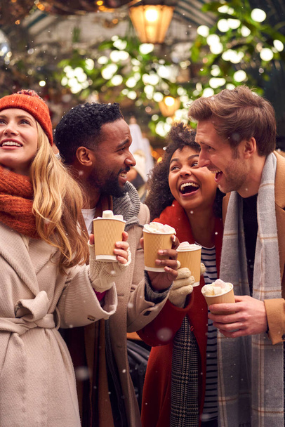 Group Of Friends Drinking Hot Chocolate With Marshmallows In Snow At Outdoor Christmas Market - 写真・画像