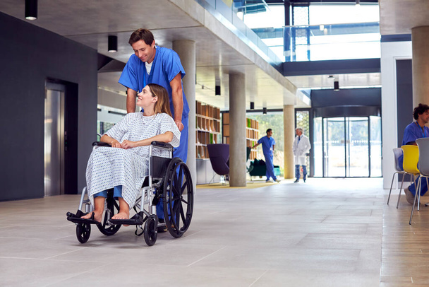 Male Nurse Wearing Scrubs Pushing Female Patient In Wheelchair Through Hospital Building - Foto, Imagem