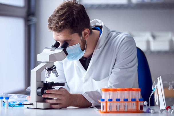 Male Lab Worker Wearing PPE Analysing Blood Samples In Laboratory With Microscope - Foto, Imagem
