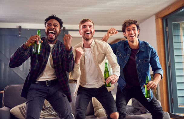 Grupo multicultural de amigos masculinos que ven juegos deportivos en la televisión en casa celebrando la meta con cerveza - Foto, Imagen