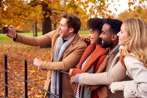 Group Of Friends Outdoors Wearing Coats And Scarves Posing For Selfie On Phone In Autumn Park - Zdjęcie, obraz