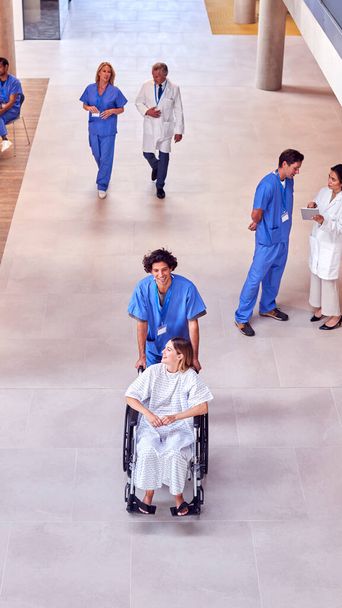 Overhead Shot Of Nurse Wearing Scrubs Pushing Female Patient In Wheelchair Through Hospital Building - Valokuva, kuva